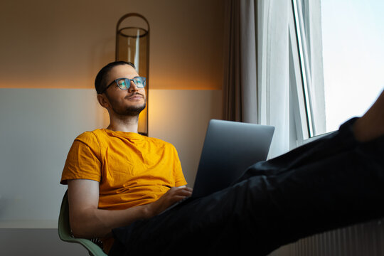 Young man working from home. Sitting on chair with laptop on legs. Wear eyeglasses and yellow shirt.