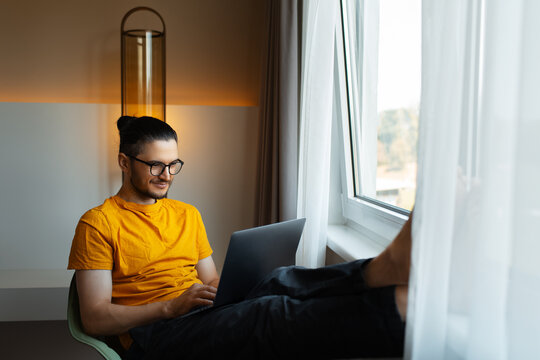 Young man working from home. Sitting on chair with laptop on legs. Wear eyeglasses and yellow shirt.