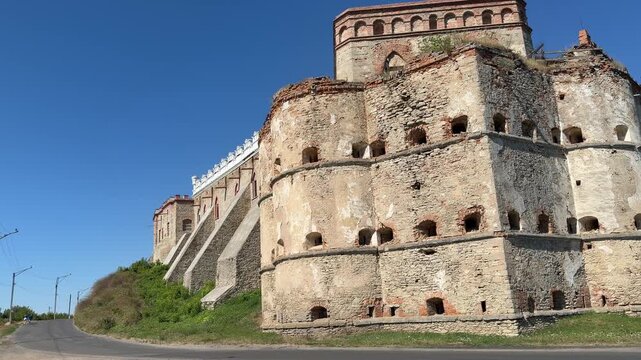 Medzhybizh Castle stone bastion with cracked walls and brick top stands beside decorative rampart, road and tree in view, Ukraine.