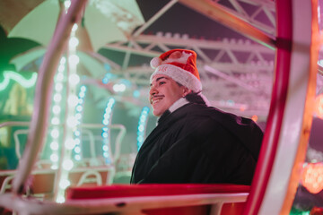 Fototapeta premium A smiling man wearing a Santa hat sits on a brightly lit amusement ride enjoying time with friends. Festive lights and colorful bokeh create a cheerful holiday atmosphere.