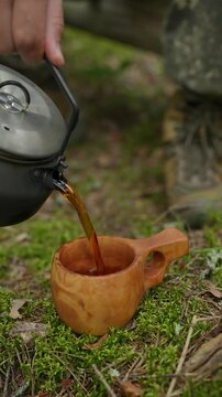 Man pouring hot dark herbal tea from a camping kettle into a traditional wooden kuksa cup on forest moss. Concept of bushcraft camp lifestyle, outdoor relaxation, wilderness survival beverage and trad