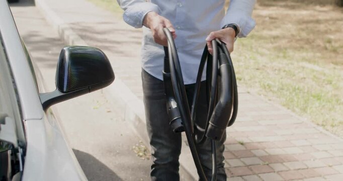 Driver of the car holds a long cable with plugs for charging an electric car and carefully twists it with his hands.