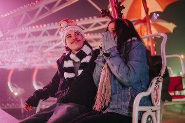 Fototapeta premium Two friends sit together on an amusement ride wearing winter clothes and holiday accessories. The pair share a cozy moment on a nighttime carnival ride surrounded by colorful lights.