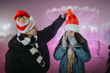 Fototapeta premium Two friends wearing Santa hats stand together at a nighttime fair. They share a playful, festive moment in winter clothing against colorful lights.