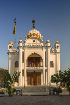 View of the white facade and golden dome of Gurdwara Shaheed Gunj Landa Bazar with a yellow flag and clear blue sky in Lahore, Punjab, Pakistan.