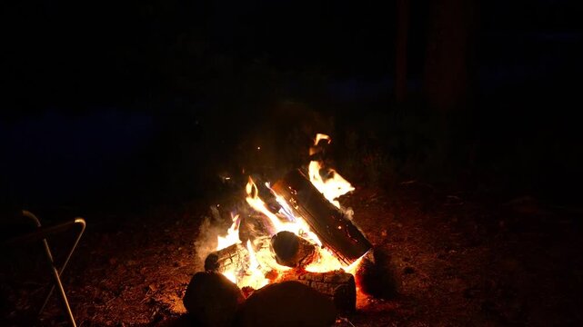 Close up of a campfire burning brightly with orange flames on wooden logs in pitch black darkness. Concept of night wild camping warmth, outdoor firecraft skills, cozy campfire atmosphere and wilderne