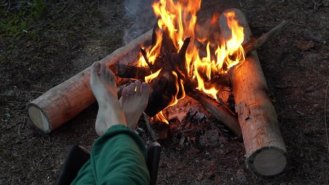 First person perspective of a person warming bare feet by a bright burning campfire in the forest. Concept of cozy outdoor relaxation, summer wild camping comfort, hiking rest break and connection wit