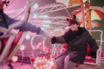 Fototapeta premium A smiling young person reaches out while sitting on a colorful amusement ride with holiday accessories. Friends share a joyful moment on a brightly lit winter carnival attraction.