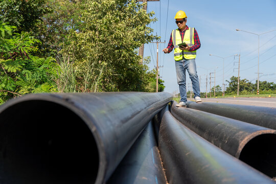 Asian Engineer inspecting large HDPE water supply pipes prepared for installation at roadside construction site, pipeline infrastructure and public utility development.