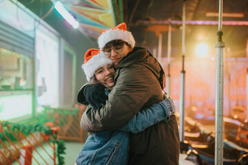 Fototapeta premium Young couple wearing Santa hats hug and smile at a holiday carnival. They embrace on a festive fairground with colorful lights and rides in the background.