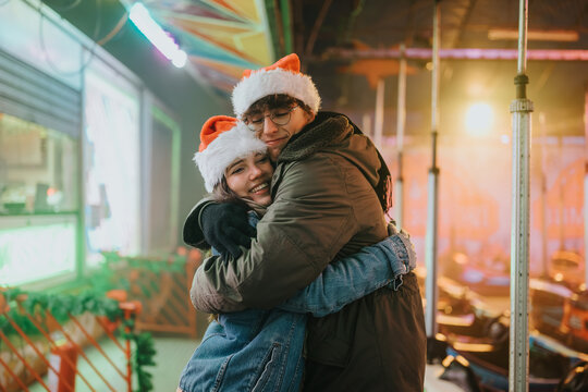 Young couple wearing Santa hats hug and smile at a holiday carnival. They embrace on a festive fairground with colorful lights and rides in the background.