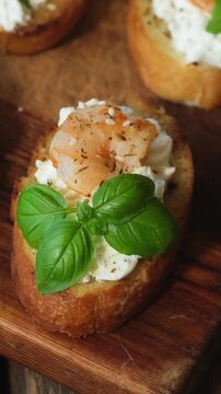 Vertical video. Bruschetta or crostini with cream cheese, shrimp and fresh basil on wooden board. Tasty snacks.