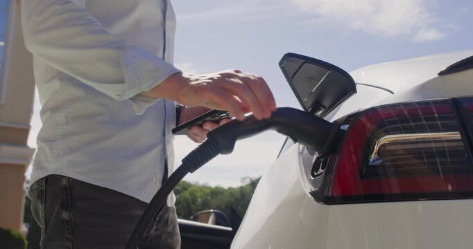 Driver uses a mobile app to remotely open the electric car's charging hatch and connects the charging station's plug to the car.