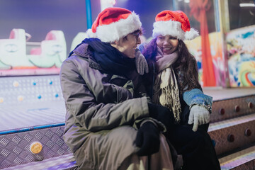 Fototapeta premium Two friends wearing Santa hats sit and smile together at an outdoor amusement ride. They share a cozy moment during a nighttime winter fair.