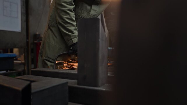 Young adult Caucasian welder grinding metal beam with angle grinder in workshop, bright sparks flying. Footage is ideal for industrial manufacturing promos and vocational training videos