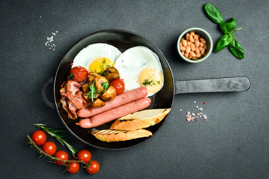Traditional American breakfast: fried eggs, sausage, bacon and vegetables in a frying pan. Homemade food. On a dark stone background.