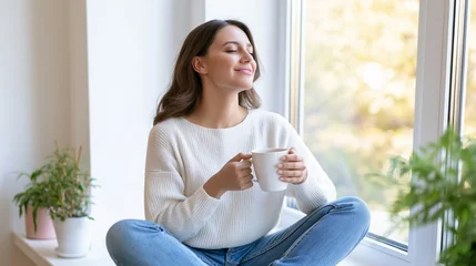 Young woman sitting cross-legged on a windowsill, holding a white mug, enjoying a moment of relaxation with sunlight filtering through the window © Nataliia
