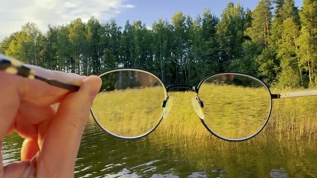 Hand Holding Eyeglasses in Front of a Calm Lake at Sunset