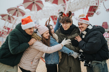 Fototapeta premium A group of friends wearing Santa hats and winter coats hug and laugh together. They enjoy a festive moment at an outdoor amusement park in cold weather.