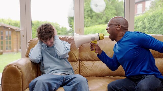 Teenagar boy staring at phone without taking eyes off it while adult man is trying to call him using megaphone. illustrating concept of the generation gap and communication problems within a family.