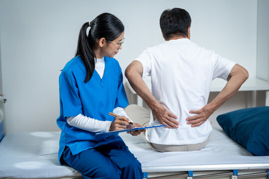 Female nurse taking notes while a man sits on a clinic bed and holds his lower back in pain