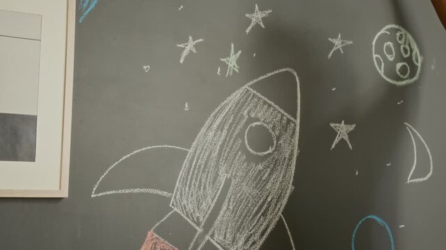 African American boy drawing rocket ship with chalk on blackboard, camera tilting up to show teenager drawing stars. Footage is ideal for educational campaigns and creative learning promotions
