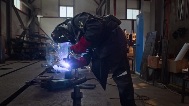 Male welder wearing protective mask and gloves welding metal structure in industrial workshop as bright sparks fly. Handheld zoom shot is ideal for manufacturing industry promos