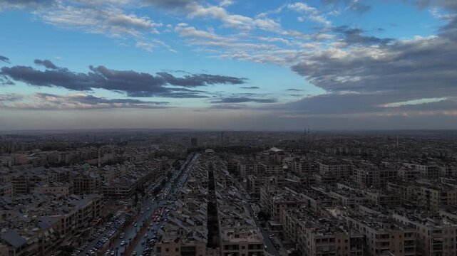 Aerial drone view of an urban landscape in Syria at sunset. City skyline and street lights during dusk. Middle Eastern city view under colorful evening clouds.