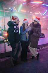 Fototapeta premium Three young friends wearing Santa hats laugh together at a neon-lit amusement ride. A winter carnival scene with bumper cars and colorful lights captures holiday fun.