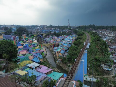 Aerial view of Jodipan, the famous colorful village with vibrant rooftops, a winding river, and a railway bridge under a cloudy sky in Blimbing, East Java, Indonesia.