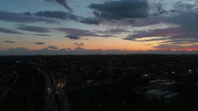 Aerial drone view of an urban landscape in Syria at sunset. City skyline and street lights during dusk. Middle Eastern city view under colorful evening clouds.