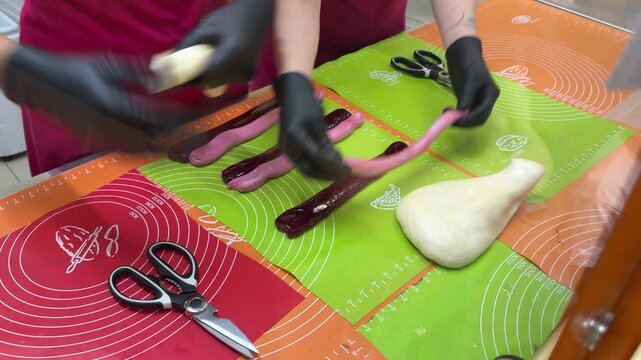 Gloved hand rolls small piece of dough beside row of dark pink candy sticks laid out on colorful mats with scissors.