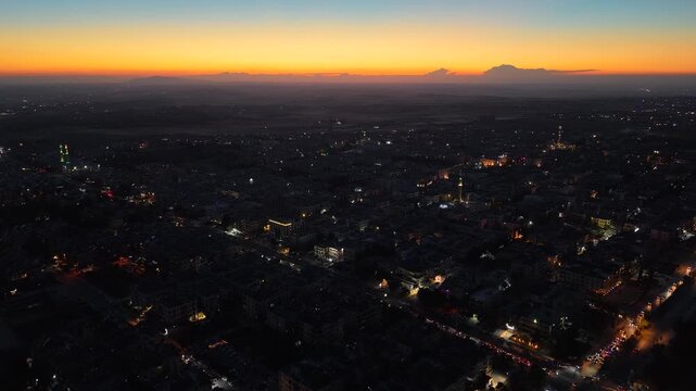 Aerial drone view of an urban landscape in Syria at sunset. City skyline and street lights during dusk. Middle Eastern city view under colorful evening clouds.