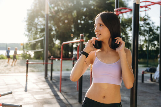 Asian female athlete wearing purple sports top adjusts headphones while standing in outdoor fitness area with exercise equipment and trees in the background