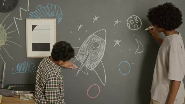 African American teenager and kid drawing rocket and moon with chalk on blackboard. Footage is ideal for STEM education marketing, creative learning campaigns, and school advertisements