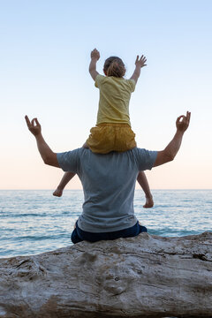 Back view of father and child enjoying sunset at the beach, relaxing together on driftwood by the sea.