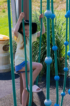 Young girl in summer clothes climbing blue rope structure at outdoor playground on a sunny day, surrounded by green grass and plants.