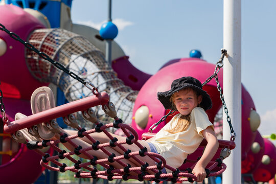 Young girl lounging on a net swing at a colorful modern playground on a bright sunny day, wearing a black sunhat and light clothing.
