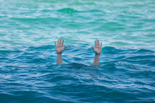 Two hands emerge from blue ocean waves, reaching upward for help, conveying urgency and safety concerns in a natural aquatic setting.