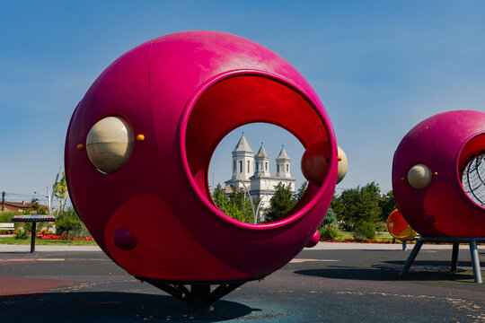 Bright pink playground structures with circular openings, set in a park, with a historic church in the background under a clear blue sky.