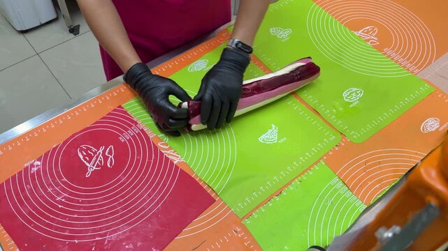Gloved hand rolls burgundy and white striped candy piece into cylindrical shape on green mat, scissors resting on red mat nearby.
