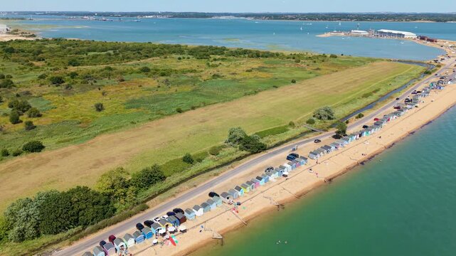 Aerial view of Calshot Beach featuring a long row of colorful beach huts along the coastline with green fields and the sea in Calshot, England, United Kingdom.