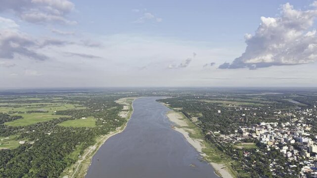A backward-moving aerial view along the river shows the contrast between Kushtia's urban landscape on one side and green natural surroundings on the other, with distant clouds above the horizon.
