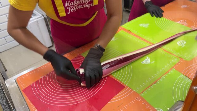 Long burgundy and white striped candy rope stretches across colorful silicone mats, gloved hands working in background of workshop.