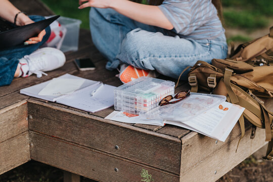 Student sitting on a wooden bench with open notebooks, art supplies and a backpack. The scene shows casual outdoor studying and creative work in a park setting.