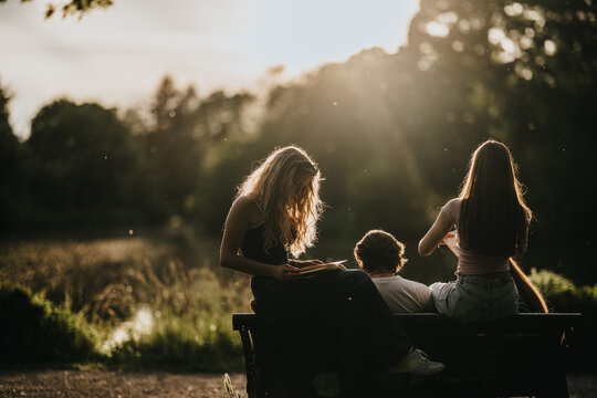 Students sit on a bench reading and relaxing in warm late-afternoon sunlight. The friends enjoy a quiet lakeside moment during golden hour outdoors.