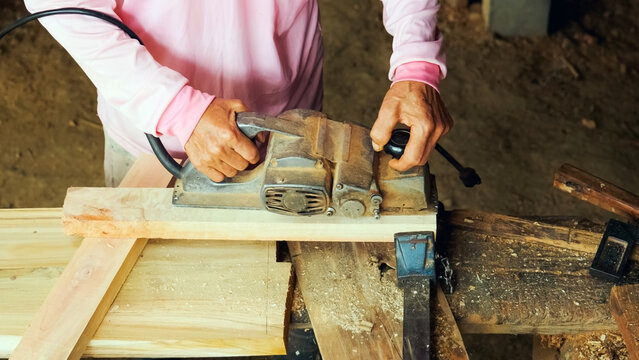 Close-up of a carpenter's hands operating an electric hand planer to shave and smooth the surface of a wooden plank in a woodworking workshop