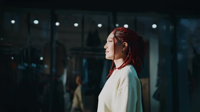 Woman with red braided hair looking thoughtfully at storefront in modern city at night