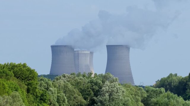 The Dampierre-en-Burly nuclear power plant in France, featuring large cooling towers emitting visible steam above a foreground of lush green trees, wide shot.
