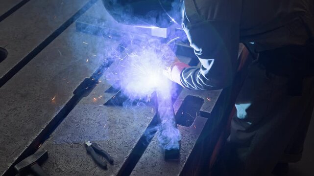 Male welder welding metal parts on workbench in workshop, bright sparks and smoke rising from joint. Footage is ideal for industrial manufacturing promos and trade school ads